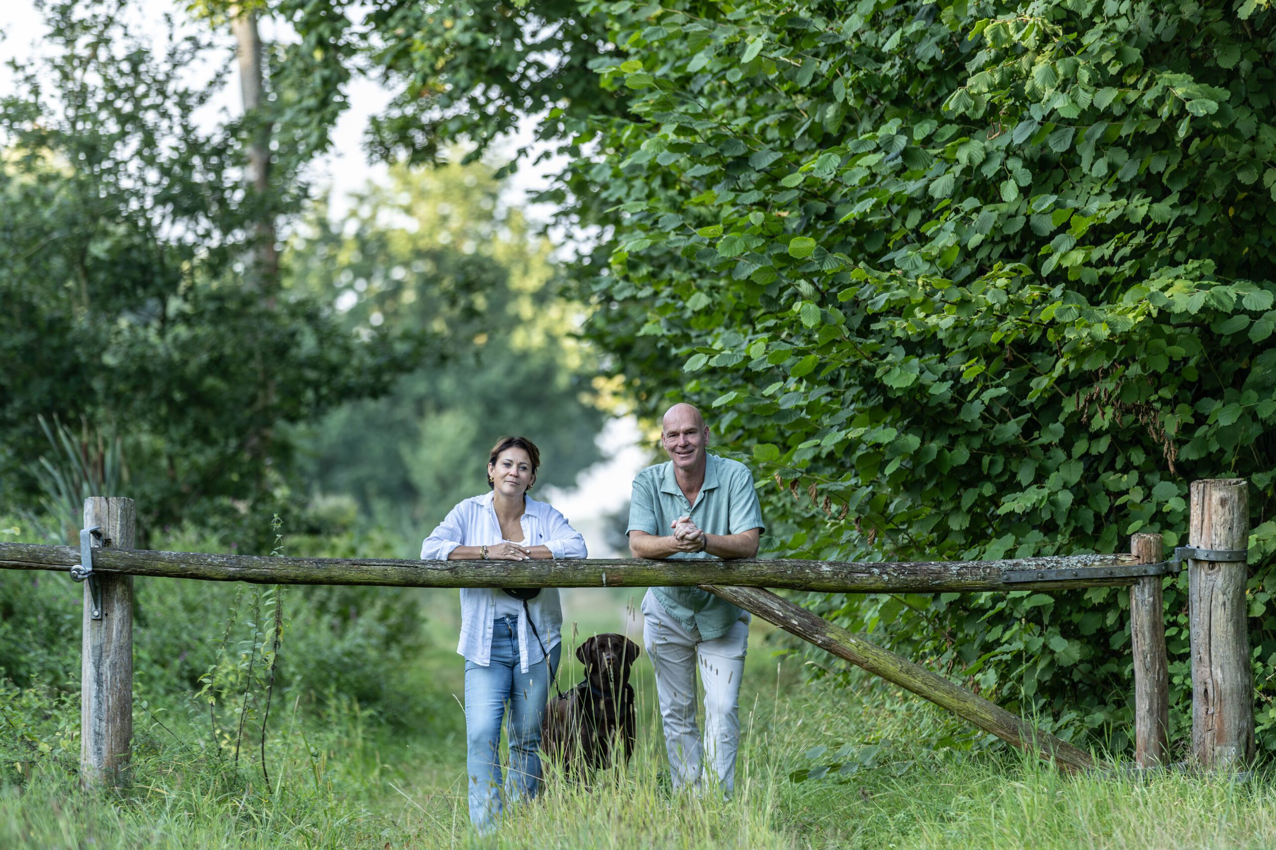 Hans en Suzanne leunen op een houten hek aan het begin van een bospad, met hun hond